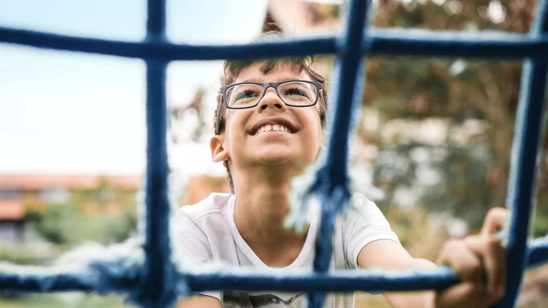 A smiling young boy in glasses looking upward and holding onto a blue net structure, with a blurred background suggesting an outdoor setting.