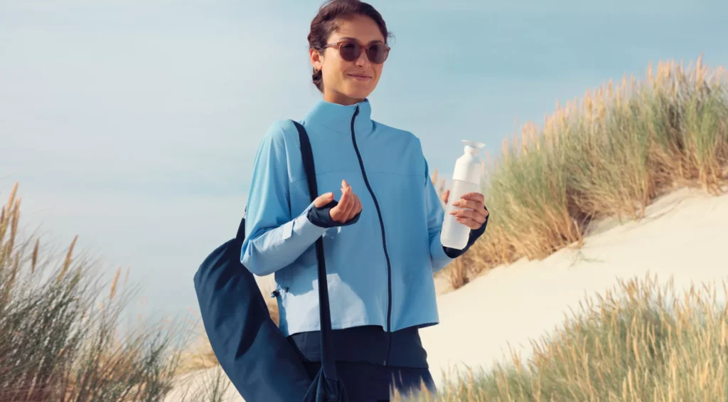 A woman in sunglasses and a blue jacket applies sunscreen on her hand while holding a bottle, with a sandy beach and dune grass in the background.
