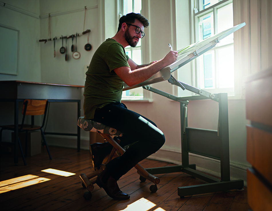 A man with glasses sketching on a drawing board in a sunlit room with wooden floors and cooking utensils hanging in the background.
