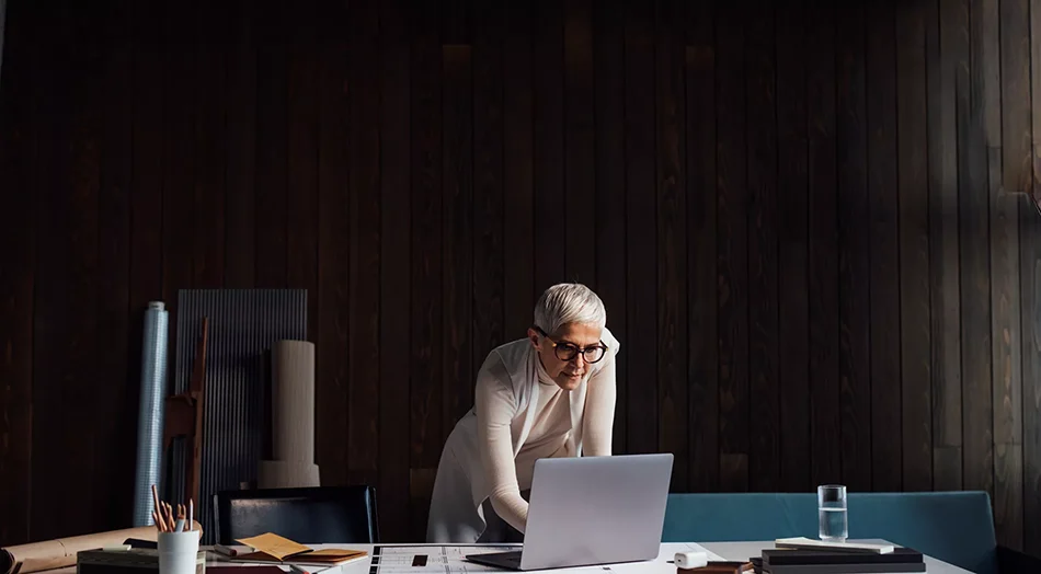 A person with short white hair and glasses, wearing a light-colored top, is concentrating on a laptop screen at a dark wooden desk with various items, including papers, a tablet, a cylindrical container, and a glass of water. They are leaning forward with their elbow on the table and their hand on their forehead, suggesting deep focus or concern. The room has a dark wooden backdrop, giving a moody atmosphere to the scene.